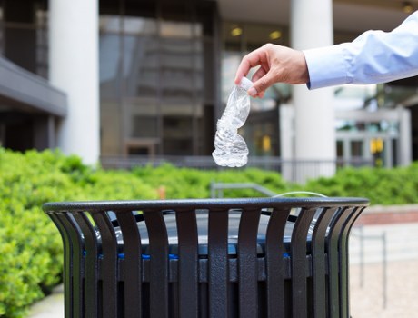 A person wearing a light blue long-sleeved shirt is disposing of a clear plastic water bottle into a black, slatted outdoor rubbish bin located on a paved area. The background shows a modern building with large windows, and there are some green shrubs and a small garden bed nearby. The scene appears to be in a residential or commercial courtyard setting, possibly within the Charlton area, with natural daylight highlighting the clean and orderly environment. The image emphasizes waste disposal in an outdoor urban space, consistent with rubbish removal services offered by Garden Clearance Charlton in the local postcode area.