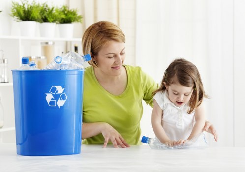 A woman with short, light brown hair and wearing a bright yellow-green top is assisting a young girl with shoulder-length brown hair, dressed in a white top, in recycling plastic bottles. They are seated at a white table in a well-lit, modern interior space with a background of white shelves filled with small potted green plants and glass containers. The woman holds a clear plastic bottle and guides the girl as she places a plastic bottle on the table. To their left, a large blue recycling bin with a white recycling symbol is filled with crushed or empty plastic bottles, some of which are transparent with blue caps. The scene conveys an environment focused on waste separation and responsible rubbish disposal, aligning with services offered by Garden Clearance Charlton in the Charlton area. The natural lighting highlights the textures of the plastic bottles and the smooth finish of the recycling bin, emphasizing environmental awareness and community engagement in waste management activities relevant to London suburbs.