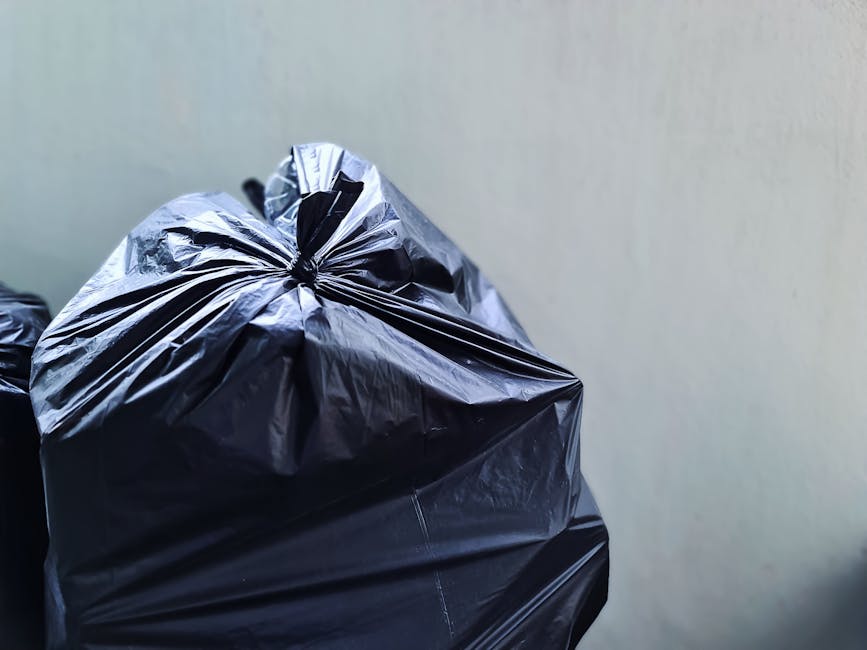 A close-up view of several black plastic rubbish bags filled and tightly knotted at the top, stacked together against a plain, light-colored wall. The bags are made of shiny, smooth plastic material a