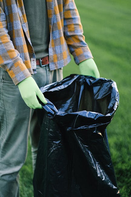 Three large black trash bags filled with waste material are placed on the pavement beside a black metal fence in an outdoor urban setting. The bags are made of thick plastic, some appearing crumpled a
